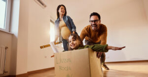 Happy expecting family having fun after moving into a new home. Father is pushing their daughter in cardboard box.