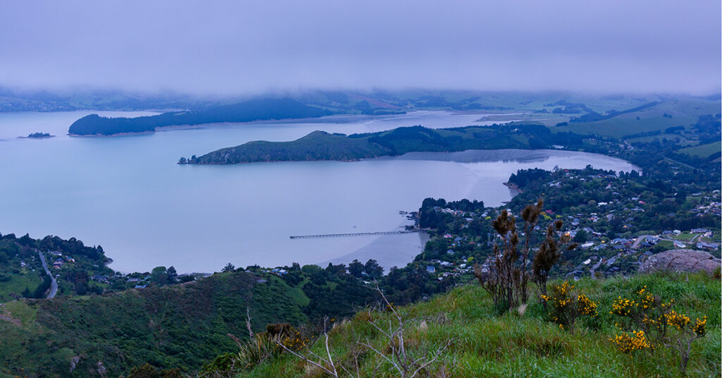 This November 2021 dusk photo shows low clouds over Te Whakaraupō Lyttelton Harbour, Aotearoa New Zealand. The community of Governors Bay is in the centre. The community jetty, with its earthquake-collapsed section, is visible. This area is part of Horomaka Banks Peninsula in Ōtautahi Christchurch.