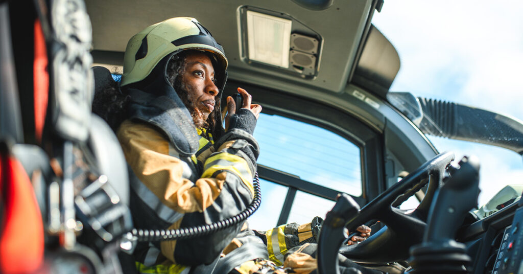 Side view of a Black female airport firefighter sitting in a fire truck using a CB station while driving towards an emergency location at the airport runway.