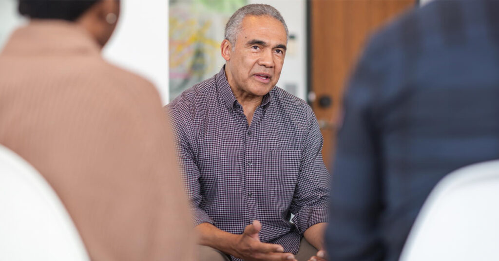 A multi-ethnic group of adults are attending a group therapy session. The attendees are seated in chairs arranged in a circle. The psychologist is a mature adult black man. He is talking to the group. He has a serious expression on his face.