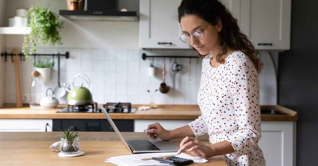 Female standing in home kitchen doing accounting work from home calculates company financial statement, use calculator fill data results on laptop application, manage family budget concept