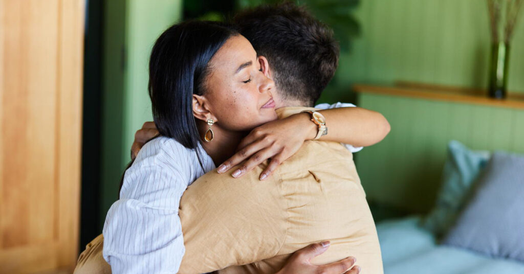 Young woman consoling her husband with a hug while sitting together on their bed at home