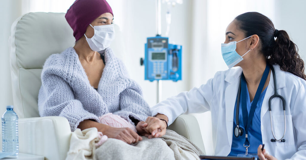 A senior woman battling cancer sits in a high back chair as she talks with her Oncologist during a Chemotherapy treatment. The patent is dressed casually in a sweater and has a blanket and headscarf on to keep her warm.