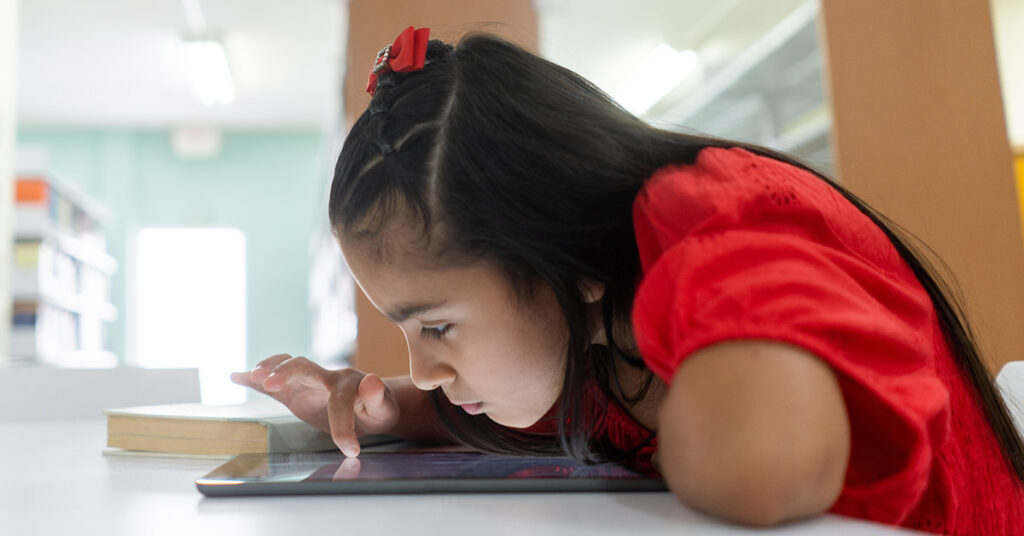 A shortsighted little latin girl using a digital tablet with her face close to the screen.