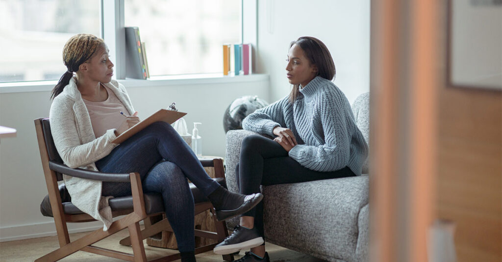 A Therapist meets with her female client in her office. The client is seated on a sofa with her arms across her body as she looks visibly nervous. The Therapist is seated in a chair in front of her as she talks about what to expect from the appointment and takes notes on her clipboard.