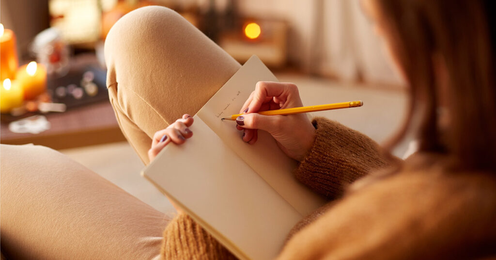 close up of young woman with pencil writing to diary and resting her feet on table at cozy home